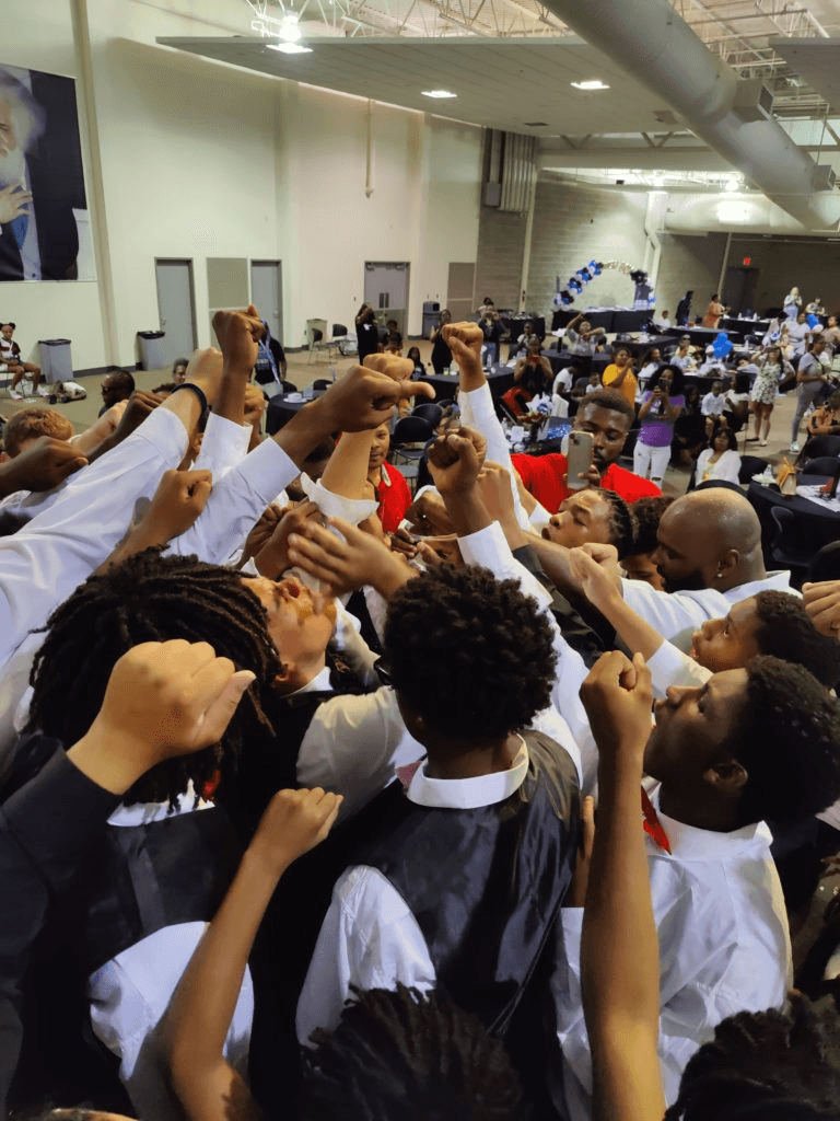Young men in white shirts and vests raise fists together in a New B.O.Y. celebration huddle